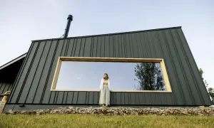 Kobieta stoi przed nowoczesnym domem pokrytym ciemną blachą na rąbek. Duże okno odbija niebo i drzewa.