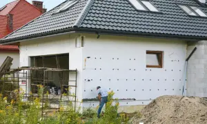 Budowa domu w toku. Izolacja ścian styropianem, widoczny fragment garażu i pracujący mężczyzna. W tle inne domy i stos piasku. Cokół domu jest jeszcze nieotynkowany.