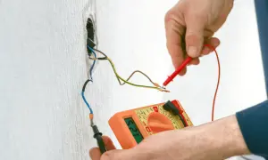 Elektryk mierzy napięcie multimetrem na przewodach instalacji elektrycznej wychodzących ze ściany.