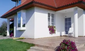 Nowoczesny dom z białym tynkiem, czerwonym dachem i charakterystycznym wykuszem. Przed wejściem kwitną fioletowe kwiaty.