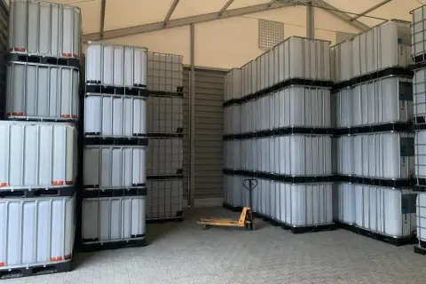 Magazyn pełen białych mauzerów 1000l, ułożonych w stosy. Widać wózek paletowy.