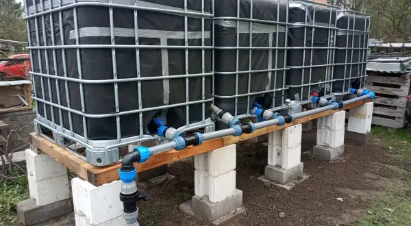 Kilka połączonych zbiorników IBC na drewnianej konstrukcji z systemem rur i zaworów.