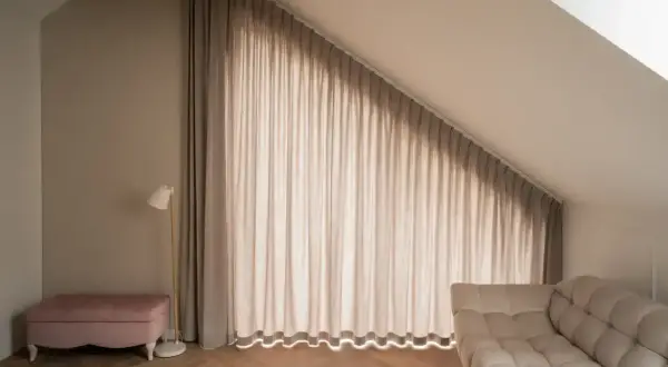 Elegancki karnisz na całą ścianę z beżowymi zasłonami, które tworzą przytulną atmosferę. Obok różowa pufa i nowoczesna lampa.