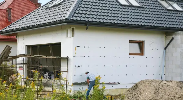 Budowa domu w toku. Izolacja ścian styropianem, widoczny fragment garażu i pracujący mężczyzna. W tle inne domy i stos piasku. Cokół domu jest jeszcze nieotynkowany.