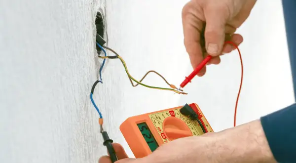 Elektryk mierzy napięcie multimetrem na przewodach instalacji elektrycznej wychodzących ze ściany.