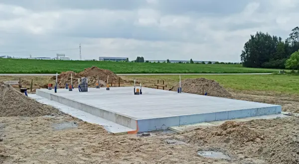 Płyta fundamentowa koszt budowy domu. Betonowa płyta na placu budowy, otoczona piaskiem i zielenią.