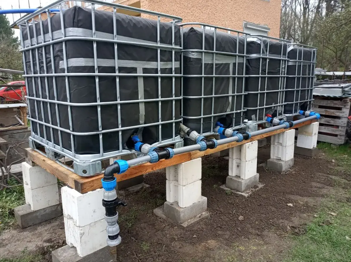 Kilka połączonych zbiorników IBC na drewnianej konstrukcji z systemem rur i zaworów.