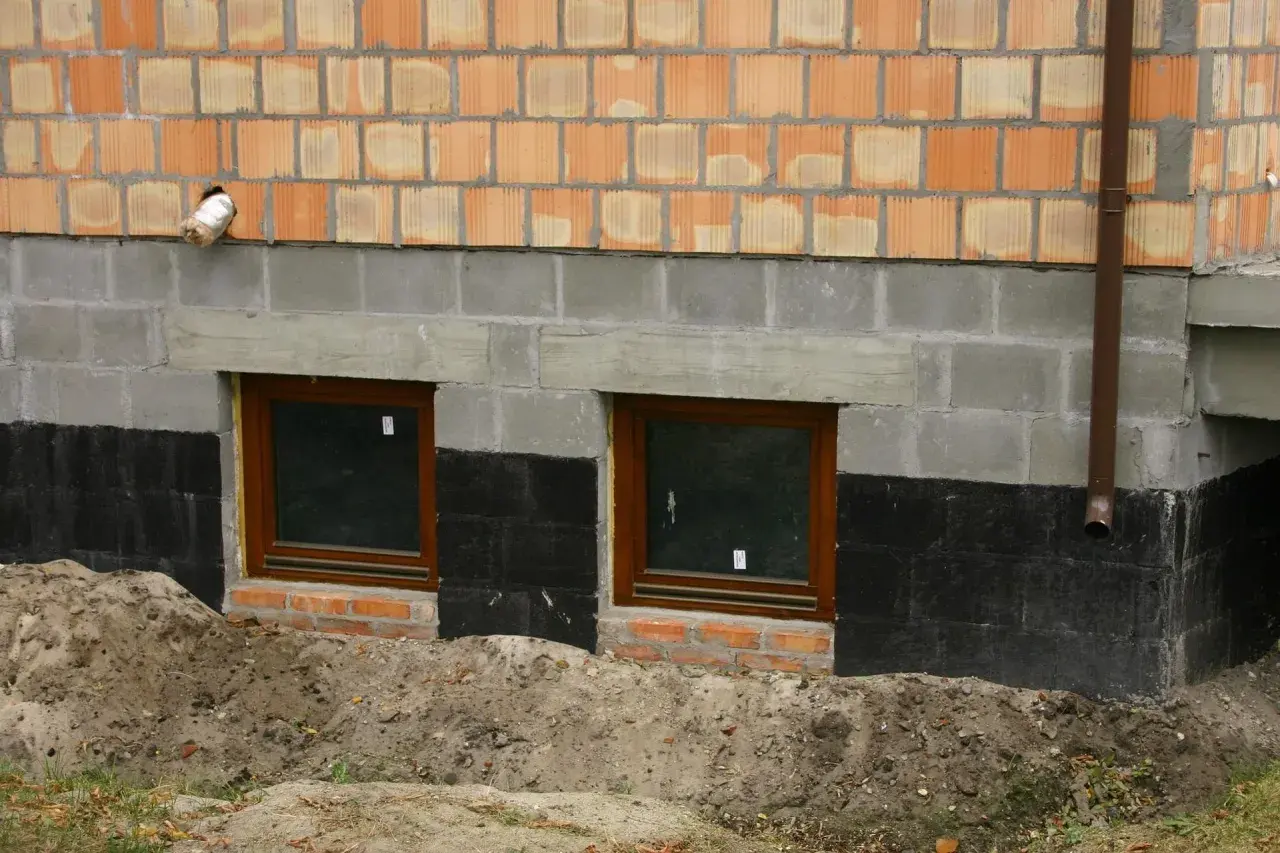 Fundament domu z oknami piwnicznymi, izolacją przeciwwilgociową i ceglanym murem.