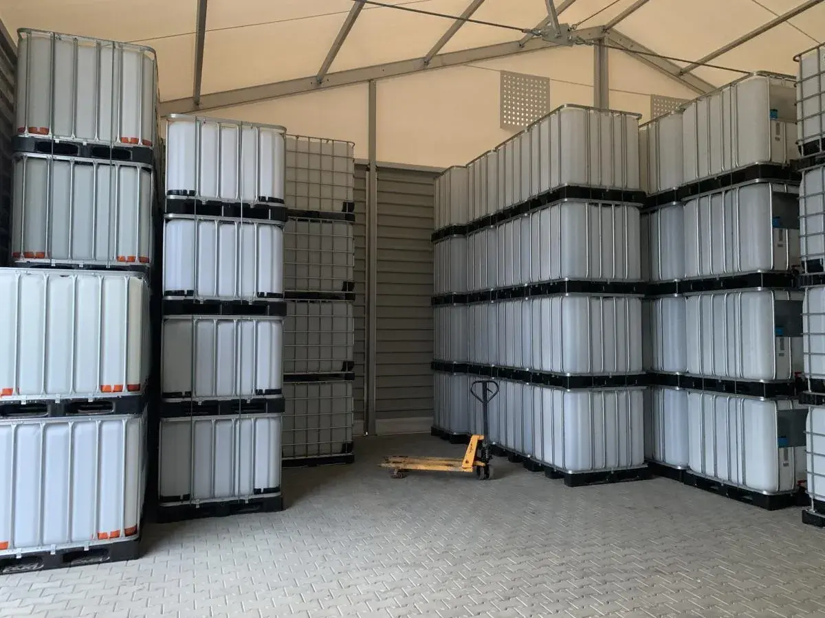 Magazyn pełen białych mauzerów 1000l, ułożonych w stosy. Widać wózek paletowy.