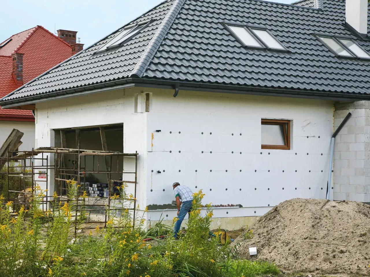 Budowa domu w toku. Izolacja ścian styropianem, widoczny fragment garażu i pracujący mężczyzna. W tle inne domy i stos piasku. Cokół domu jest jeszcze nieotynkowany.
