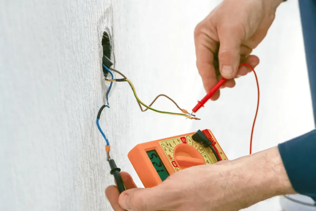Elektryk mierzy napięcie multimetrem na przewodach instalacji elektrycznej wychodzących ze ściany.