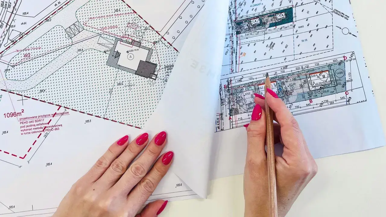 Rysunek techniczny z zaznaczonymi przyłączami i budynkiem. Kobieta z pomalowanymi paznokciami analizuje projekt, zastanawiając się, ile kosztuje pozwolenie na budowę.
