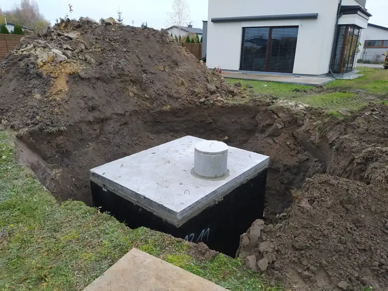 Zbiornik na wodę opadową w wykopie, obok domu w budowie. Koparka usypała kopiec ziemi.