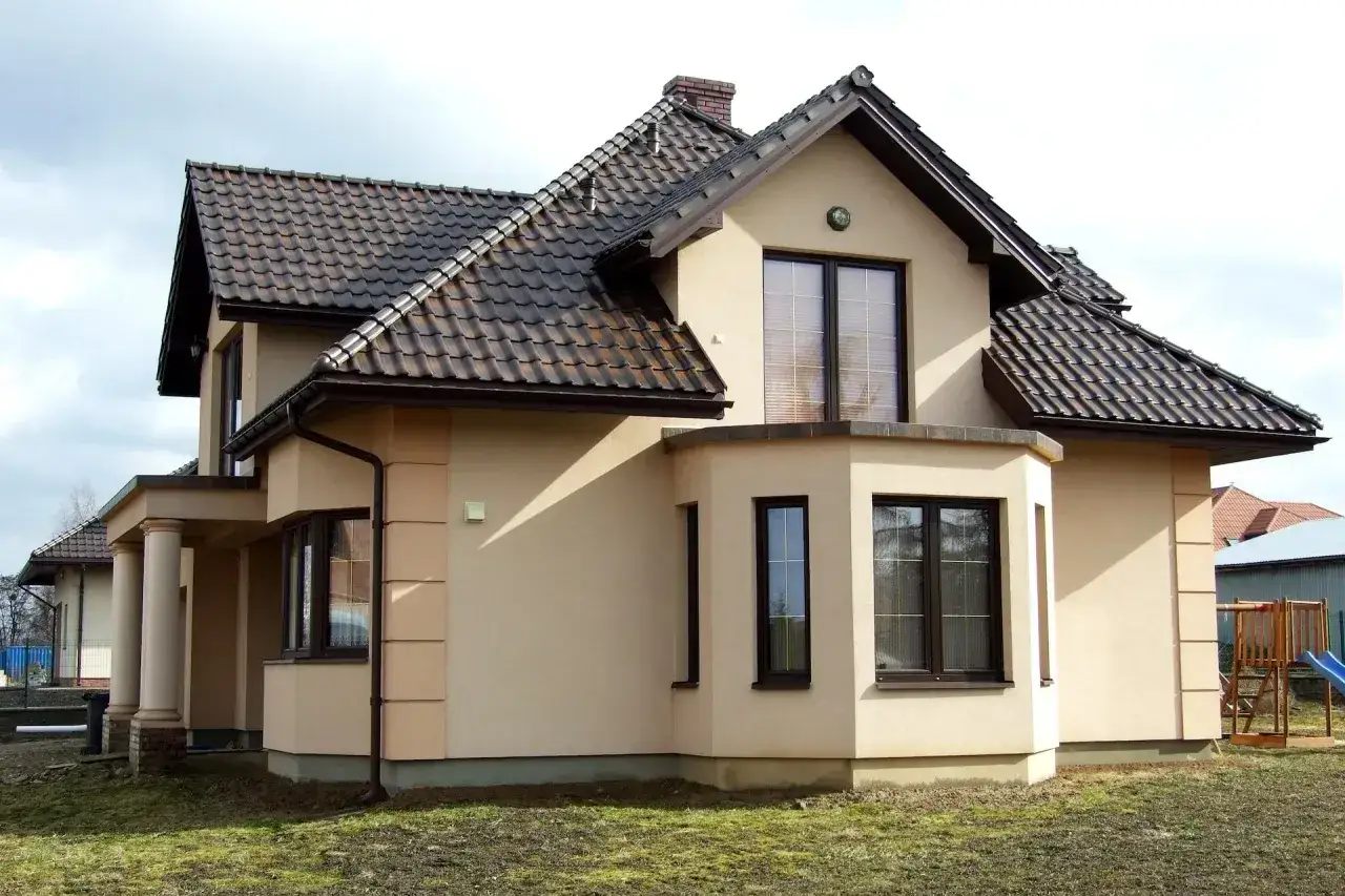 Elewacja domu w kolorze kawa z mlekiem, z brązowym dachem i ciemnymi oknami. W tle plac zabaw.