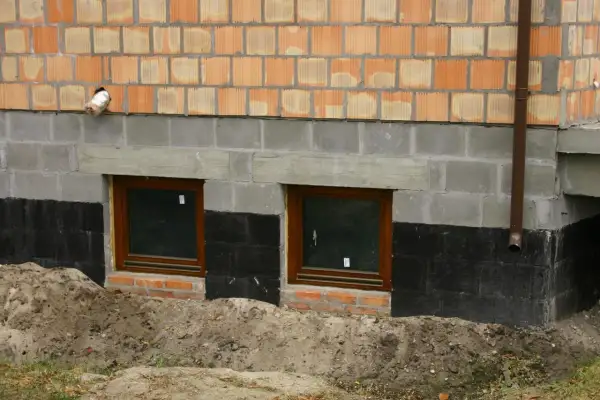 Fundament domu z oknami piwnicznymi, izolacją przeciwwilgociową i ceglanym murem.