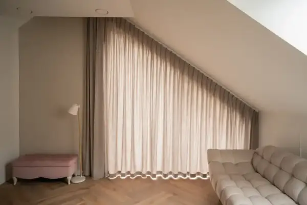Elegancki karnisz na całą ścianę z beżowymi zasłonami, które tworzą przytulną atmosferę. Obok różowa pufa i nowoczesna lampa.