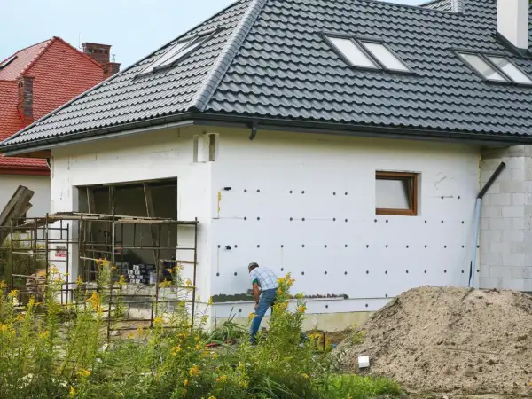 Budowa domu w toku. Izolacja ścian styropianem, widoczny fragment garażu i pracujący mężczyzna. W tle inne domy i stos piasku. Cokół domu jest jeszcze nieotynkowany.