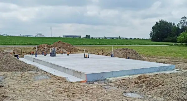 Płyta fundamentowa koszt budowy domu. Betonowa płyta na placu budowy, otoczona piaskiem i zielenią.